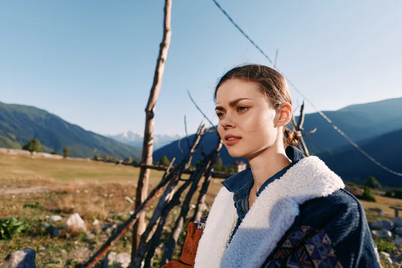 Woman portrait in countryside by a wooden fence with mountains in the background, outdoor rural scene at golden hour. Young female looking thoughtful amid nature and sunlight