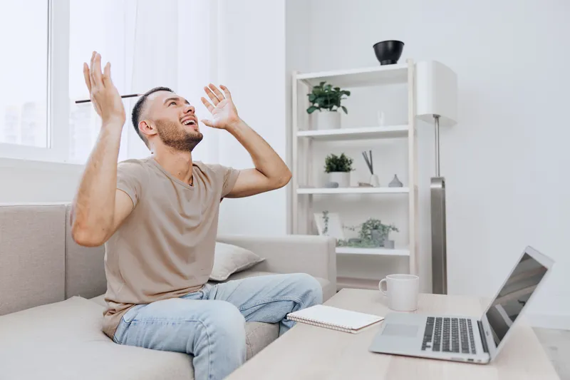 A frustrated man sitting on a sofa, expressing his emotions with raised arms, surrounded by a modern living space. Ideal for themes of stress and work-life balance.