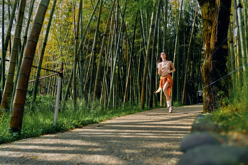 Woman jogging on a bamboo forest path for fitness, runner training in nature on a scenic trail, active outdoor exercise with shorts and jacket tied, focused cardio workout and wellness.