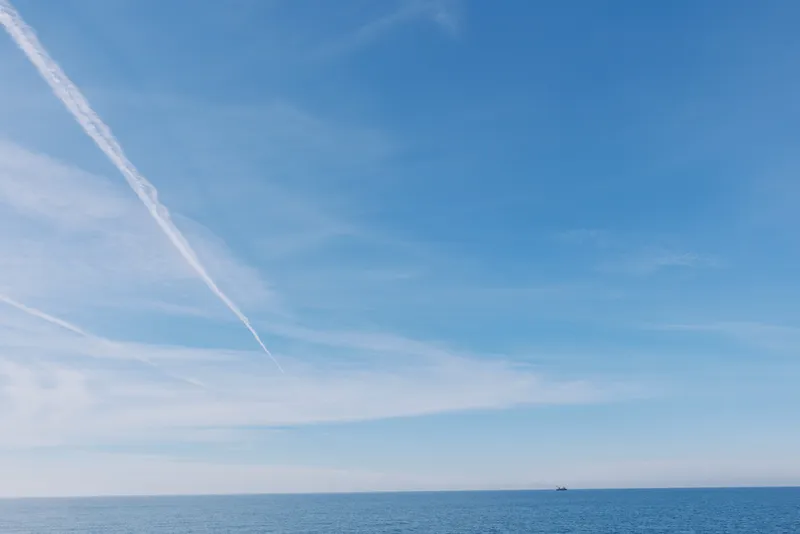 Blue sky over calm ocean with a distant boat on the horizon, white clouds and a prominent contrail creating a serene seascape