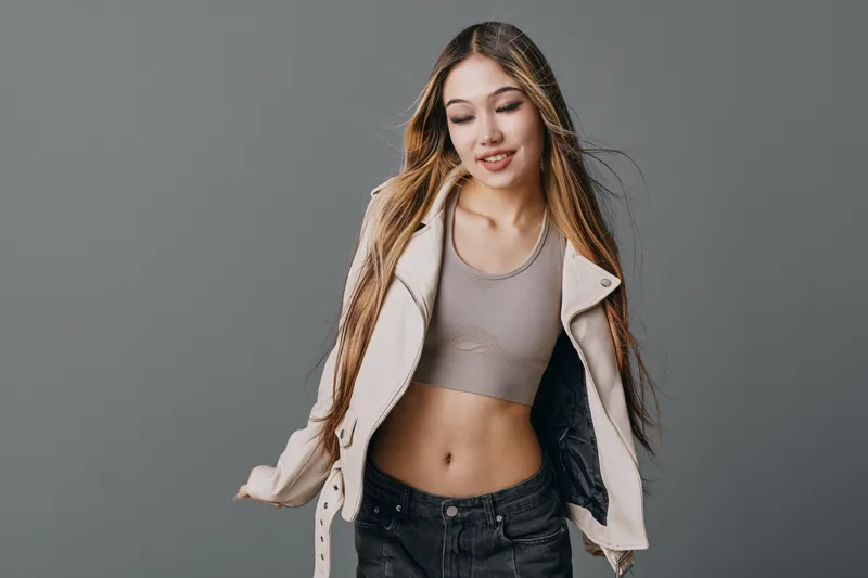 Young woman with long hair wearing casual crop top and jacket posing with eyes closed in studio. Model shows relaxed mood and fashion style against gray background.