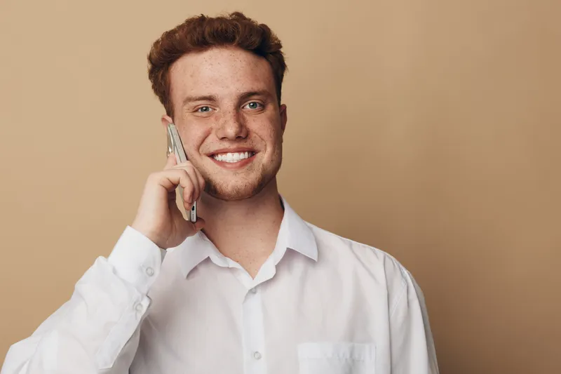 Smiling young man with curly hair wearing white shirt talks on smartphone against beige background. Concept of communication, technology, positive emotions, and casual lifestyle portrait.