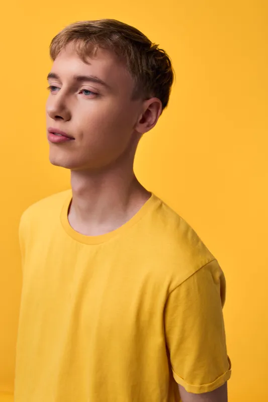 Yellow shirted male model with calm expression stands against a matching yellow backdrop, soft lighting highlights color harmony and minimalist studio composition