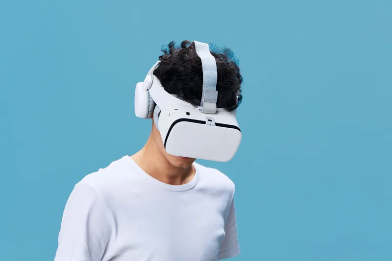 Young man wearing white VR headset exploring virtual reality against a solid blue background, embracing futuristic technology and immersive experience.
