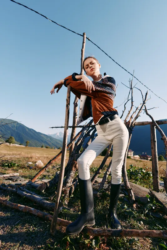 Woman model in countryside wearing sweater and rubber boots, striking a confident pose while leaning on rustic fence with mountains and open field in natural sunlight.