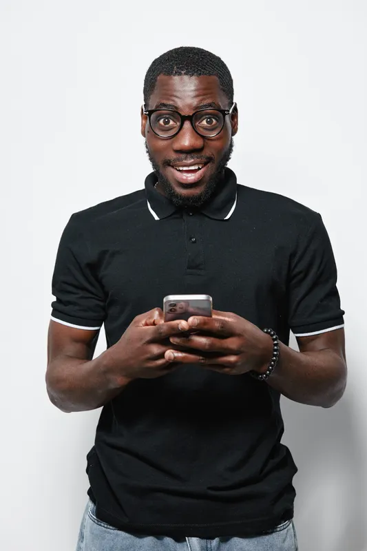 African American man with dark skin wearing glasses and black polo shirt holding smartphone and smiling while standing against white background in casual style portrait.
