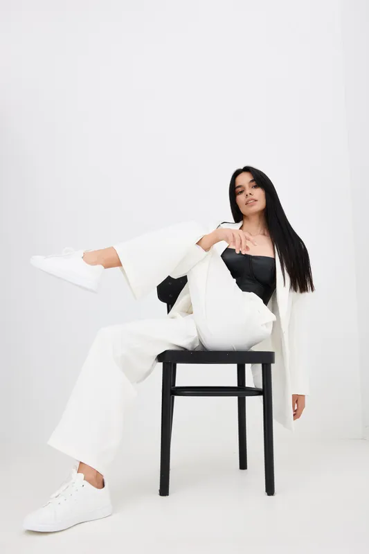 Young woman with long dark hair wearing white suit and sneakers poses confidently sitting on black chair against white background in modern minimalistic style.