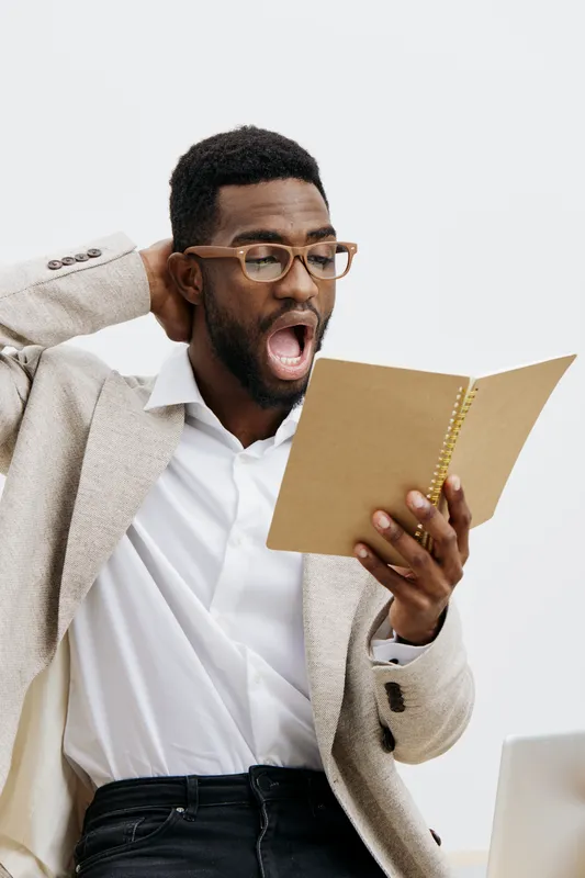 Confused man wearing glasses and beige blazer holding a notebook, looking surprised and scratching his head, isolated on plain white background, studio portrait. People lifestyle concept, office, emot