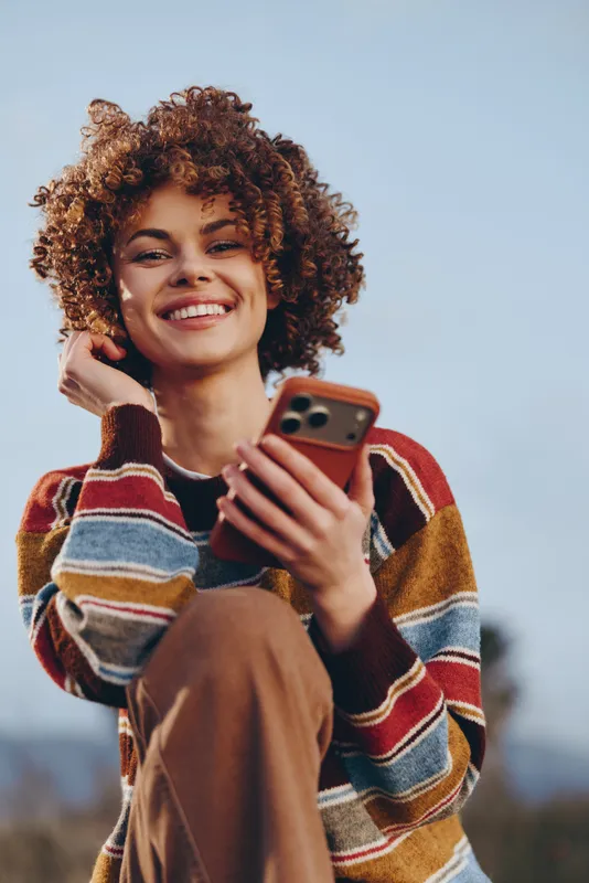 Woman with curly hair, smiling, holding smartphone outdoors, wearing a striped sweater. Warm sunlight and a soft sky background show casual lifestyle, relaxed joy, and everyday connection.