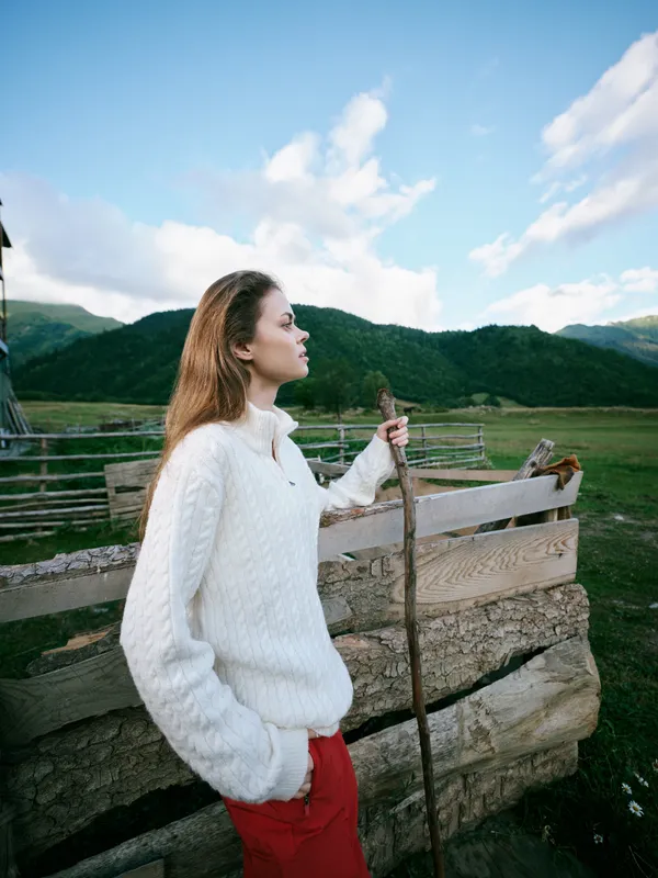 Woman in a white knit sweater leans on a wooden staff in a rural field with a rustic fence, green hills and blue sky, a calm outdoor exploration moment