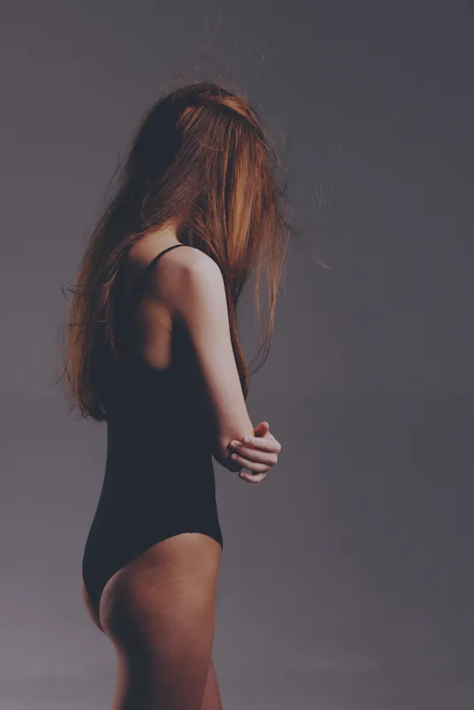 woman with long hair in black swimsuit posing with arms crossed against plain background