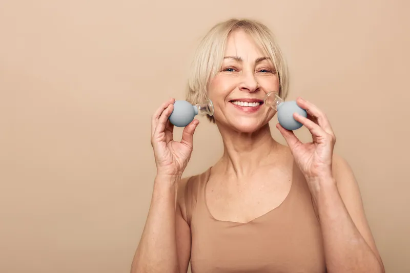 Smiling mature woman with blonde hair holds two blue facial massage balls near her cheeks. Neutral background and natural skin emphasize wellness, skincare, and self-care routine for aging skin.