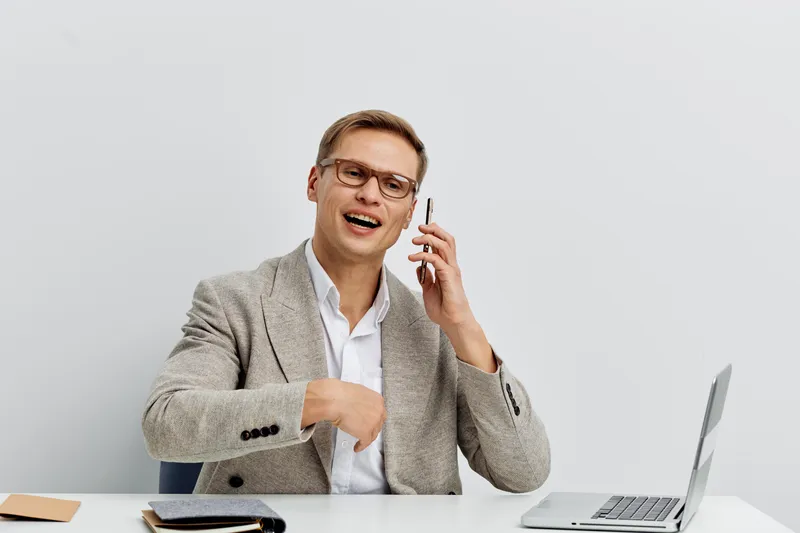 Smiling man in beige blazer talking on mobile phone while sitting at a desk with laptop and notepad, casual office environment, professional business attire, cheerful mood, modern workspace. People li