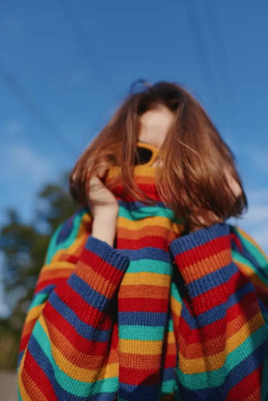 Young woman in her early 20s with red hair and sunglasses pulling a colorful striped sweater up over her face, playful and shy portrait outdoors under a clear blue sky, lifestyle fashion.