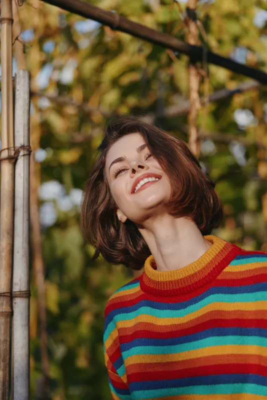 Young woman smiling woman in a colorful striped sweater, short brown hair, outdoor portrait with warm natural light, joyful expression and casual outfit leaning on a wooden pole, lifestyle.