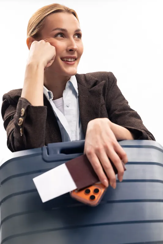Smiling woman with suitcase holding passport and smartphone looks away thoughtfully. Travel concept with luggage, vacation, and planning on white background. Positive mood.