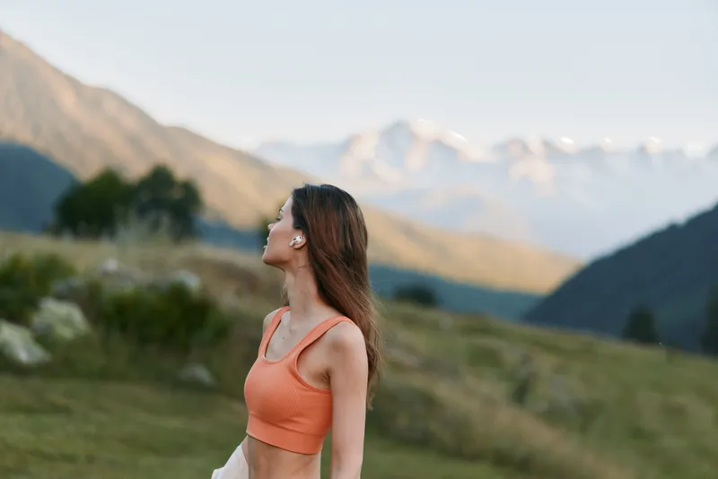 A cheerful young woman stands outdoors in a scenic mountain landscape, wearing an orange crop top. She gazes upward with a relaxed smile, enjoying fresh air and nature.