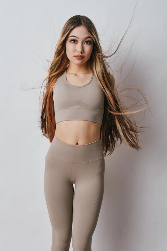 Young woman with long hair wearing beige fitness outfit posing against white background, looking confident and healthy with natural makeup and casual style.