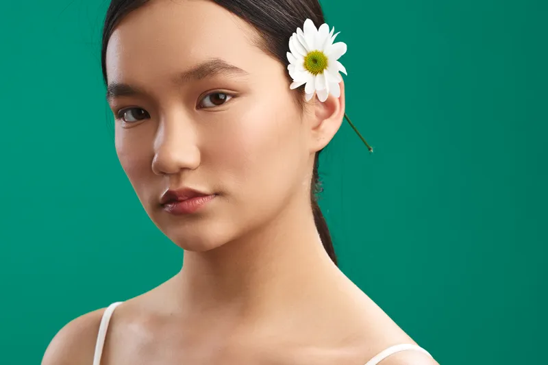 Asian woman with a white flower tucked behind her ear gazes softly at the camera against a vivid green backdrop, exuding calm elegance and natural beauty.