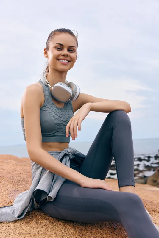 Smiling young woman in sporty attire sitting on a rock by the sea, embodying a healthy lifestyle, with a soft color palette and a relaxed atmosphere