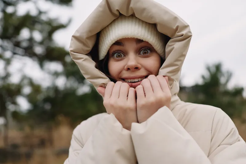 Winter scene with a smiling woman in a beige coat and knit beanie, hands near her face, creating a warm, hopeful mood outdoors in the cold air for lifestyle fashion or travel use.