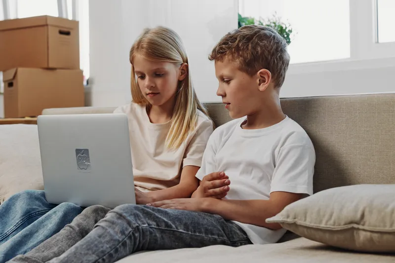 Two children sitting on a sofa using a laptop together in a bright room. The girl and boy focus on the screen, surrounded by cardboard boxes. Concept of technology, learning, and teamwork.