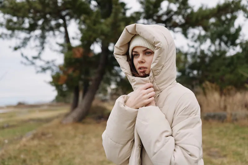 Winter outdoor portrait of a thoughtful person in a hooded coat adjusting the collar in windy scenery with bare trees and soft muted landscape.
