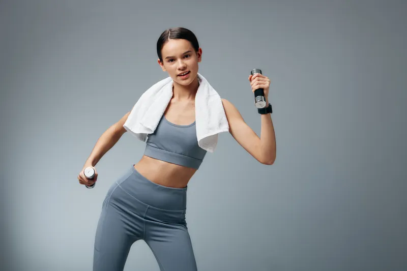Fitness woman in a gray athletic outfit lifts dumbbells with a towel draped over shoulders, mid motion in a clean studio for sport, workout and strength training.