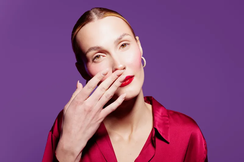 Portrait of a woman with red lipstick and hoop earrings wearing a red satin shirt on purple background. She touches her face with her hand, expressing elegance and fashion style.