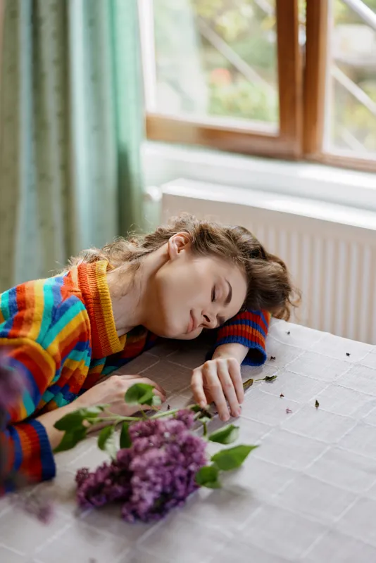 Young girl resting on a table indoors, colorful sweater, soft daylight, lilac bouquet nearby, peaceful nap moment, cozy home atmosphere, gentle breeze