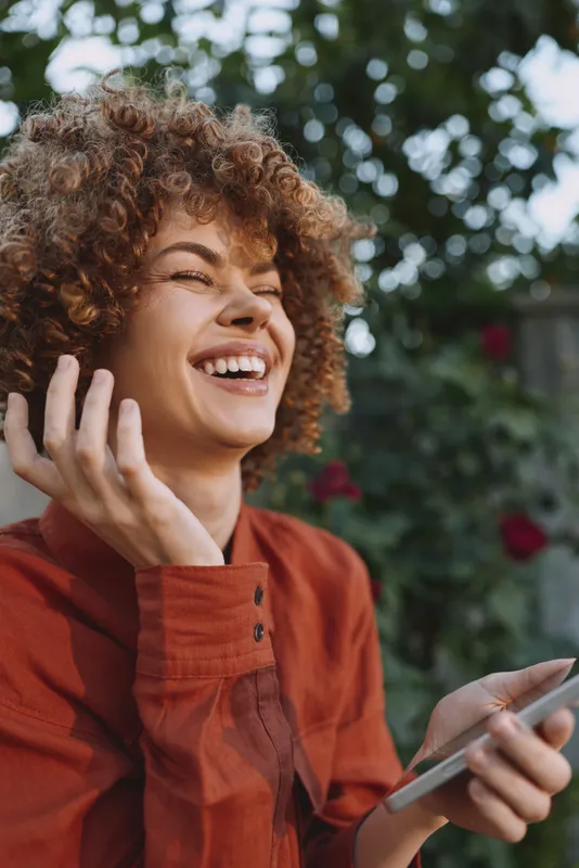 A young woman with curly hair laughs while holding a smartphone in an outdoor garden, radiating genuine joy, relaxed confidence and a casual lifestyle under leafy shade