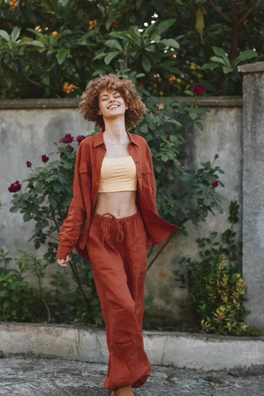 Woman in rust jacket and matching pants, cropped top, smiling, lifestyle portrait outdoors, relaxed street vibe with greenery and a concrete wall