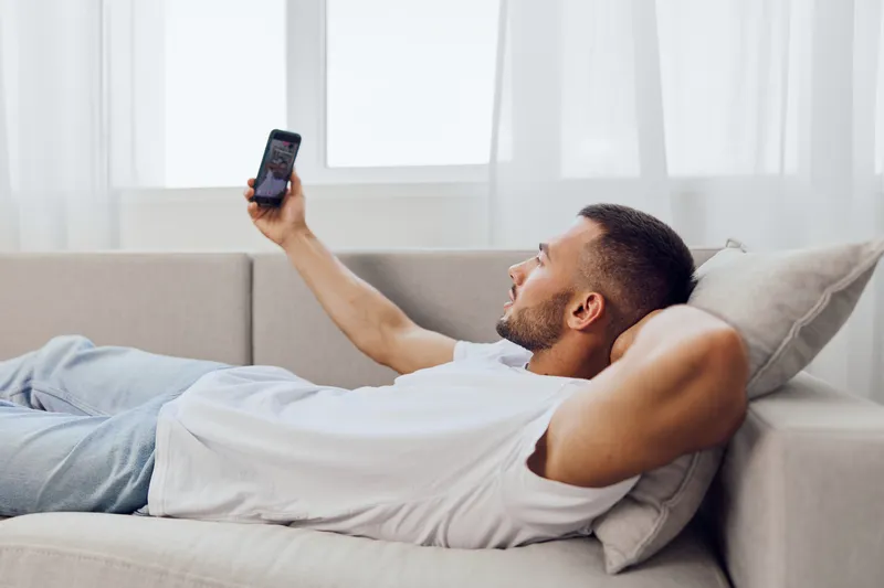 Relaxed man enjoying smartphone in cozy living room, wearing casual attire and smiling while reclining on the sofa. A serene atmosphere complements his leisure time.