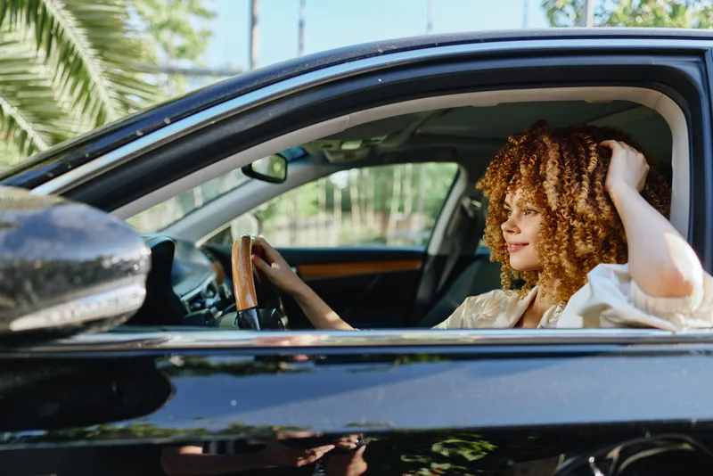 young woman driving black car with relaxed expression in sunny environment, natural light highlighting her curly hair and casual outfit
