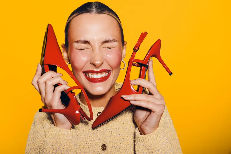 Happy woman with closed eyes holds red high heels near face on yellow background. Concept of fashion, style, footwear, and joyful emotions in studio lighting.