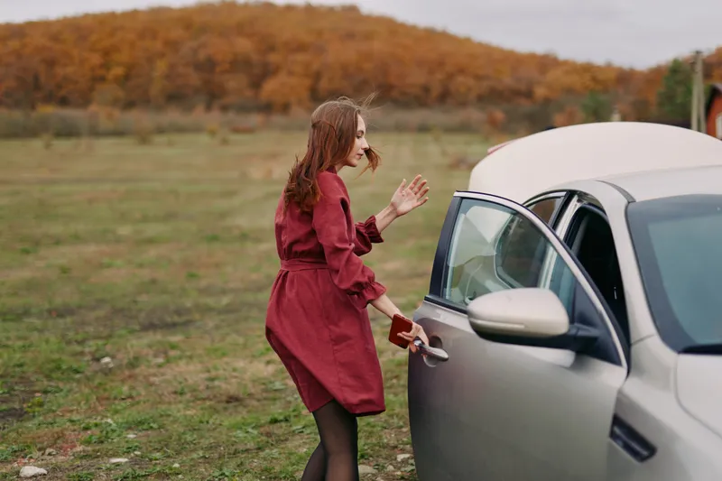 A candid autumn scene shows a woman in a red dress stepping toward an open car trunk, conveying authenticity through informal posture, genuine expression, and natural lighting.