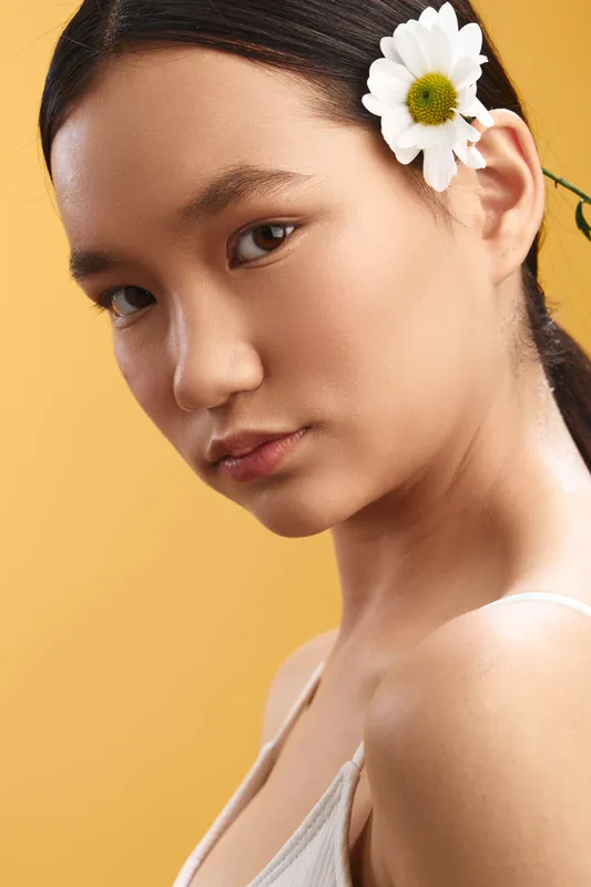asian woman with a flower tucked behind her ear, soft lighting and calm expression, portrait in a warm studio tone, conveying elegance and poise in a minimal, graceful setting.