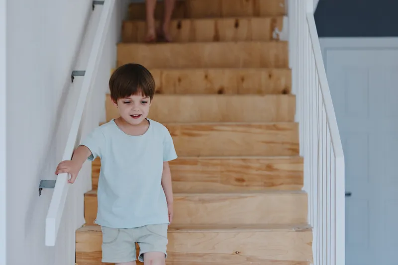 Happy boy walking down wooden stairs, bright and cheerful atmosphere, wearing light blue t-shirt and shorts, playful expression, home interior.