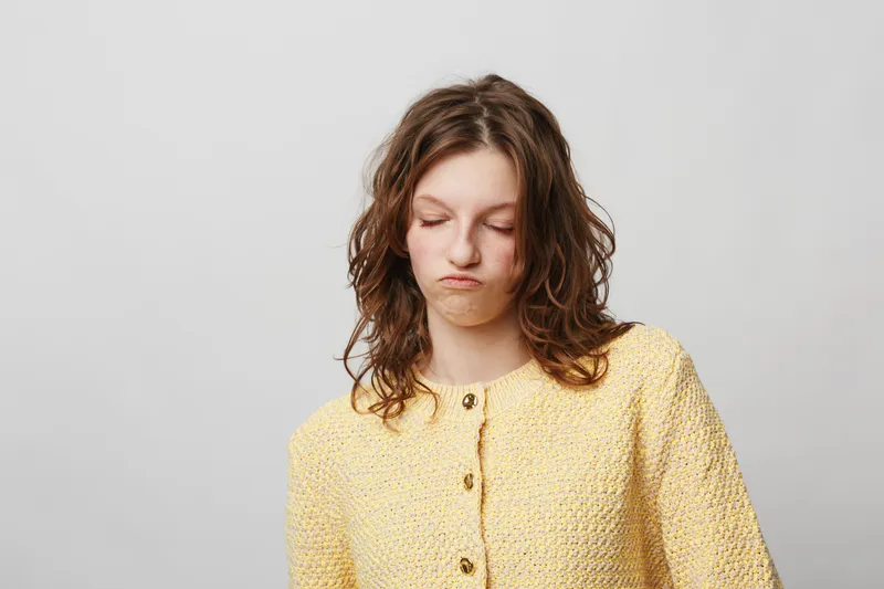 Young woman with curly hair wearing yellow jacket looks disappointed with closed eyes and pursed lips. Isolated on grey background. Concept of dissatisfaction, frustration, and mood.