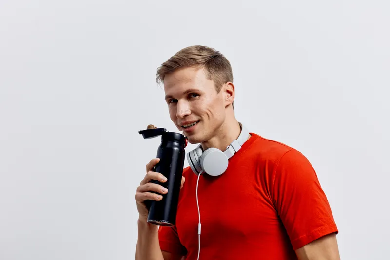 Smiling man wearing red t-shirt holding black thermos with white headphones around neck isolated on plain white background studio portrait cheerful lifestyle concept