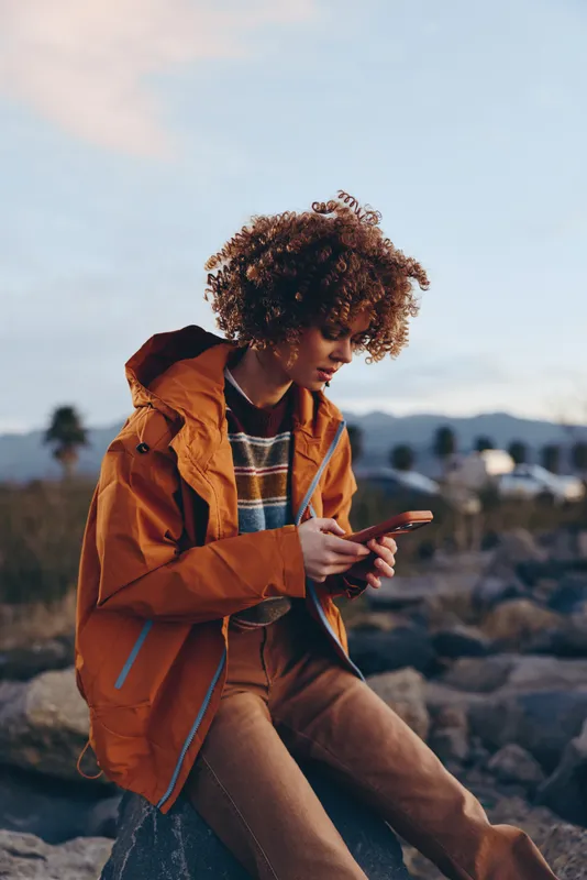 Woman lifestyle image, young woman with curly hair, orange jacket, sitting on rocks outdoors near mountains, using smartphone and looking at screen, golden evening sky, blurred background