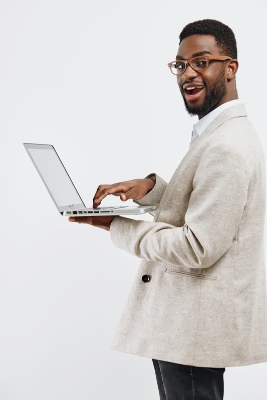 Smiling man with beard wearing glasses and beige blazer holding laptop computer, looking at camera, happy expression, casual business attire, indoor studio shot, white background. People lifestyle con