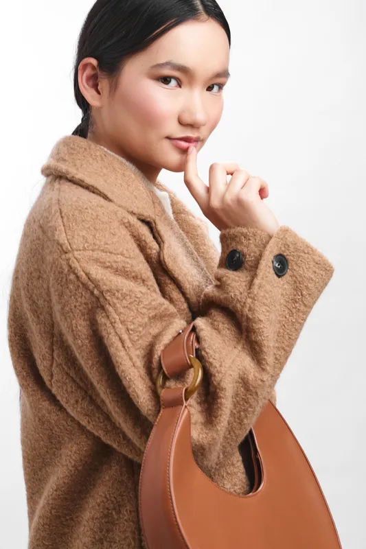 asian woman wearing a cozy brown coat holds a stylish leather bag, presenting a chic autumn look in a clean studio setting with warm tones and soft lighting.