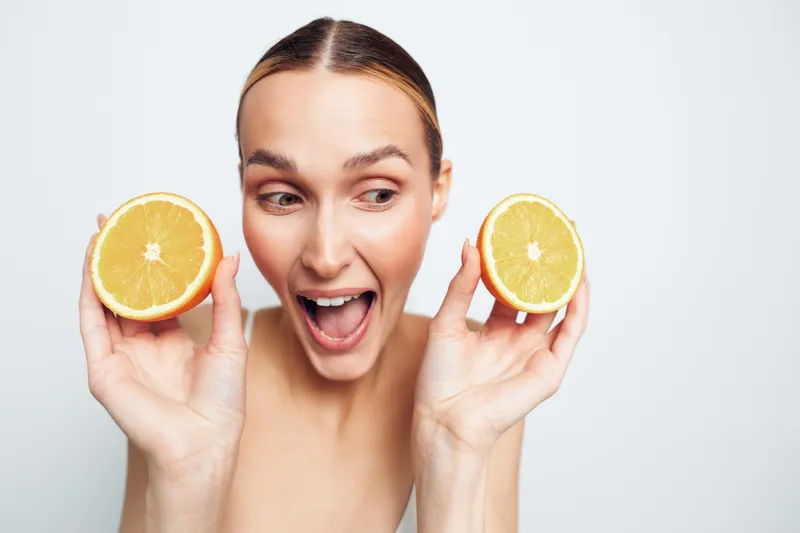 Young woman holds two halves of fresh orange near face looks excited and happy isolated on white background with natural skin and no makeup.
