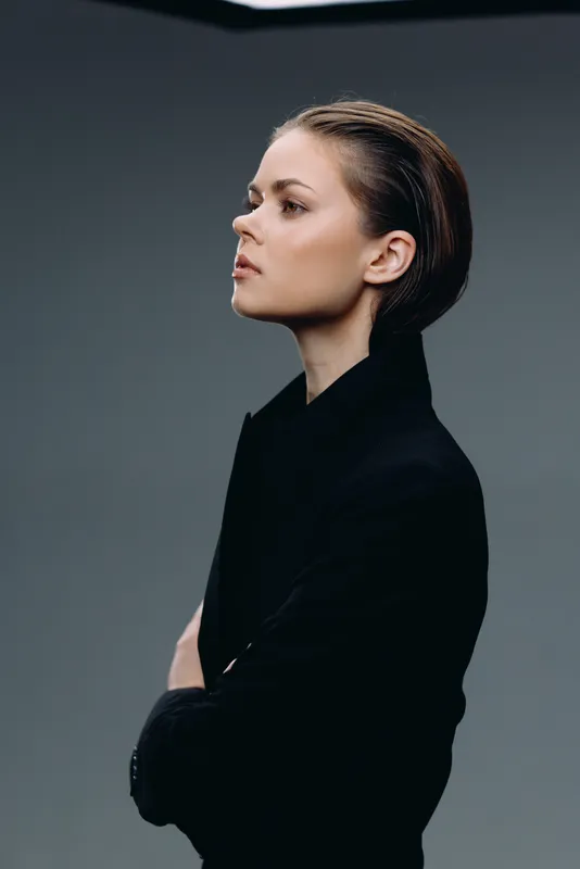 Profile of a confident woman with braided hair wearing a black blazer, posing against a gray background in a studio setting.
