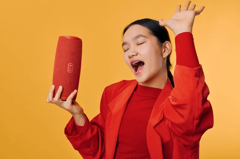 Young woman in red jacket and sweater holding portable speaker, singing with closed eyes and raised hand on yellow background for music and entertainment concept.