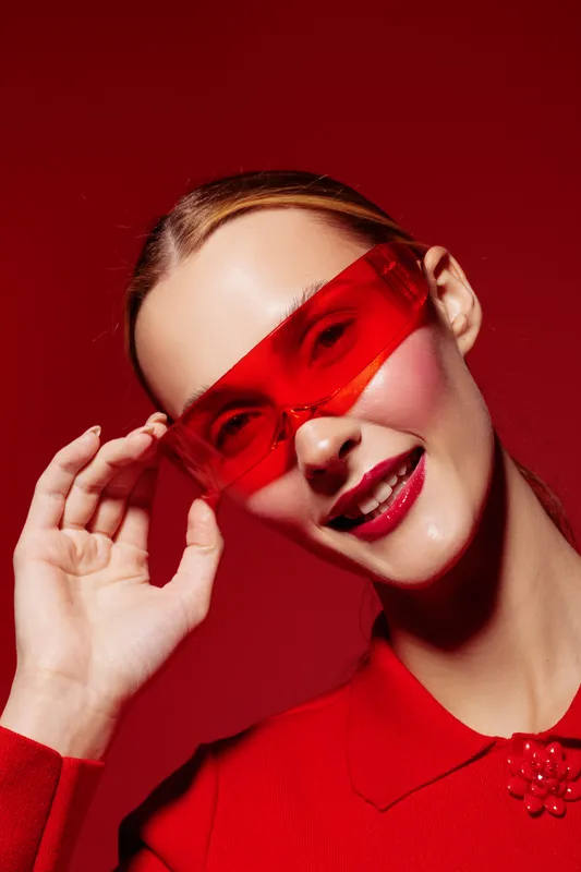 Smiling woman with red sunglasses and red outfit poses against red background. Fashion portrait with glossy lips and sleek hairstyle. Concept of style, confidence, and modern beauty.