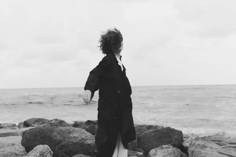 Child on rocky shoreline wearing a flowing coat, reaching toward the sea as wind sweeps the horizon, evoking solitude, exploration and outdoor resilience.
