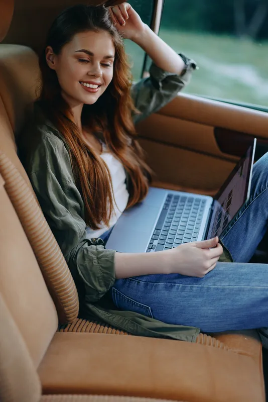 Young woman enjoying her time working on a laptop in a stylish car interior, showcasing a relaxed and cheerful expression perfect for digital nomads.
