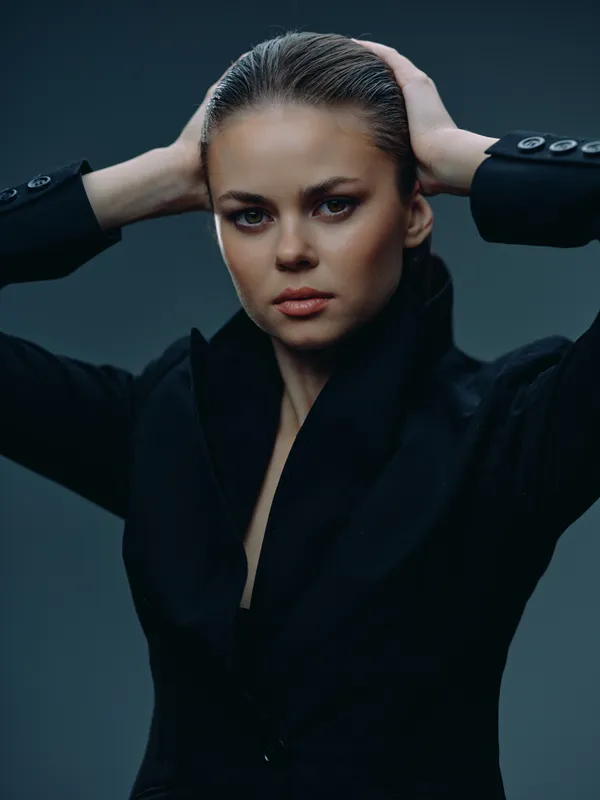 Serious woman with slicked-back hair wearing a black blazer posing confidently with hands behind head under moody lighting